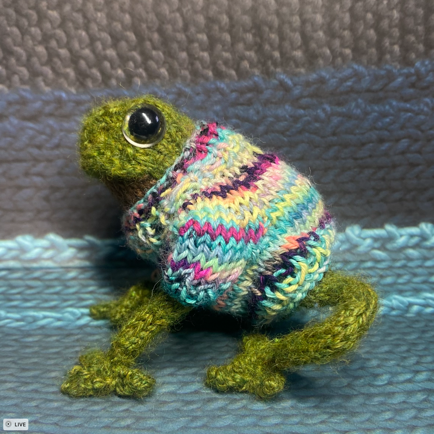 A green knit toy frog is wearing a multi-coloured knit sweater featuring bright and vibrant colours. The frog is sitting sideways on a background that is a mix of grey, dark blue, and light blue.