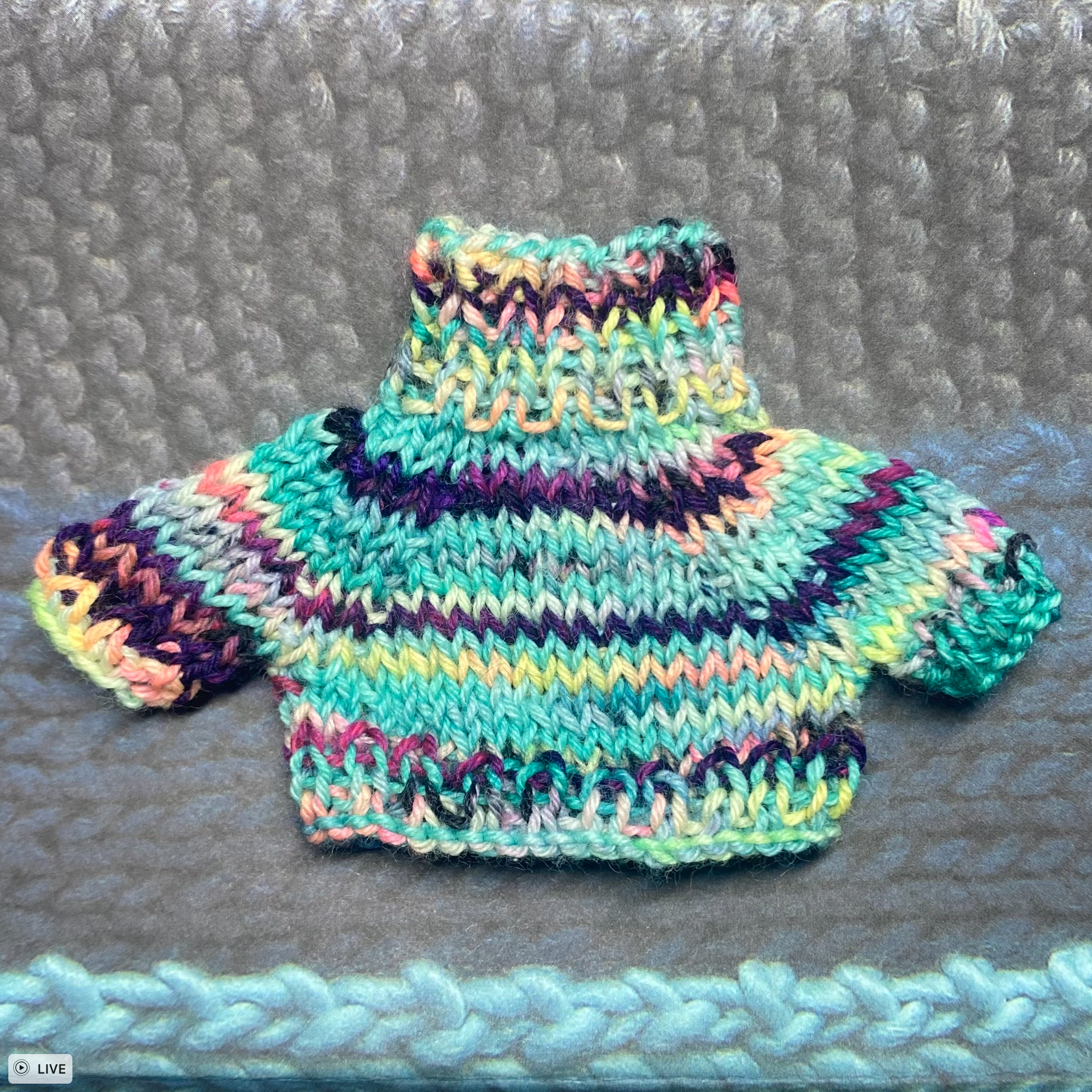 A multi-coloured knit sweater for a toy frog is shown from the front on a background that features shades of grey, dark blue, and light blue. The sweater is brightly coloured with shades of yellow, green, blue, orange, pink, and purple.