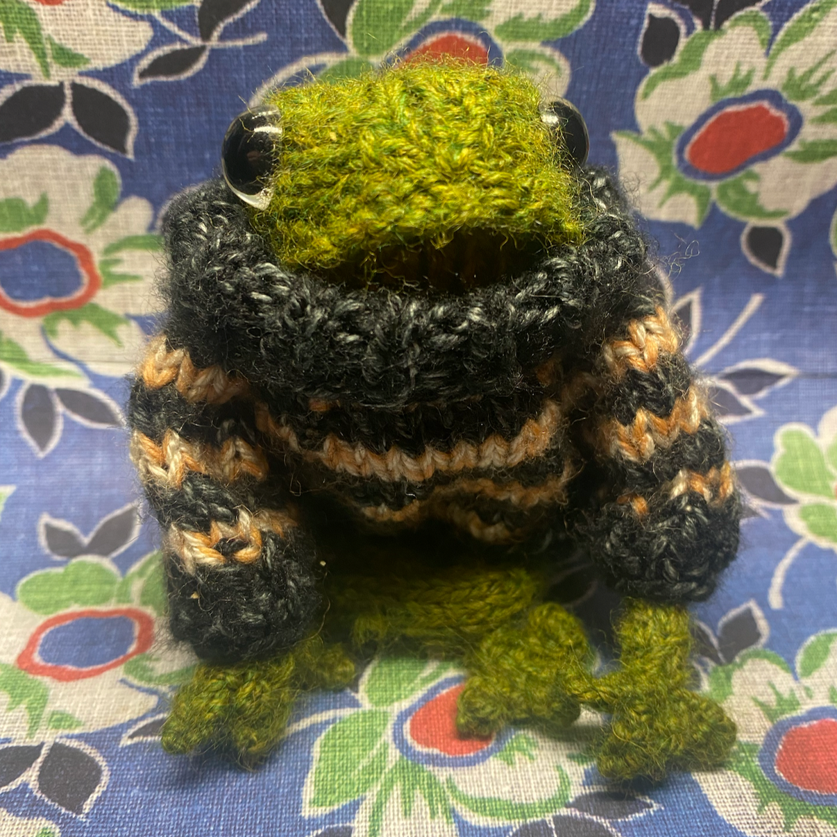 A cute knit frog named Trevor sits on a blue background decorated with green, white, and red flowers. Trevor is wearing a black and orange knitted sweater with the neck folded down, adding a charming and cozy touch to the cheerful setting.