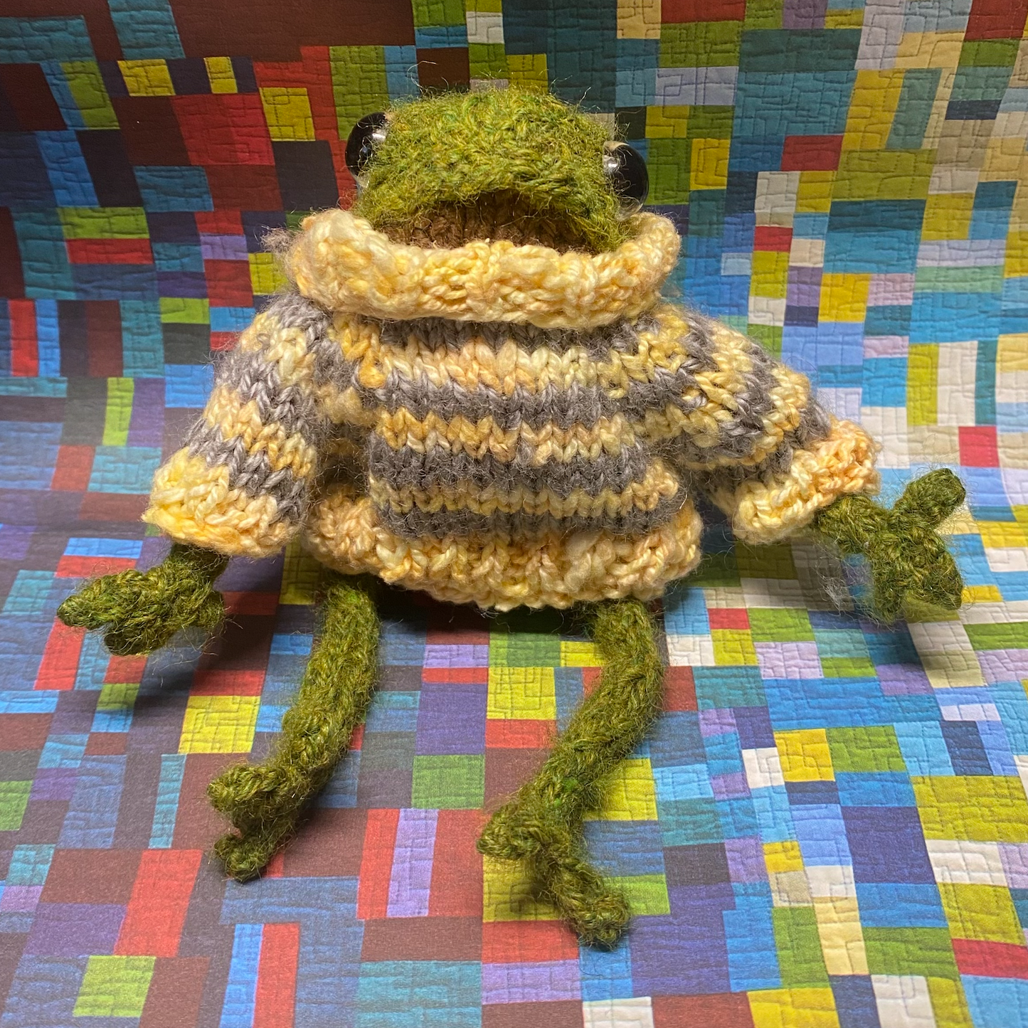 Trevor the toy frog is green with a brown belly and sits regally on a paper multicoloured quilted background. Trevor is wearing a tiny yet fashionable yellow and grey striped sweater and looks extra cozy.