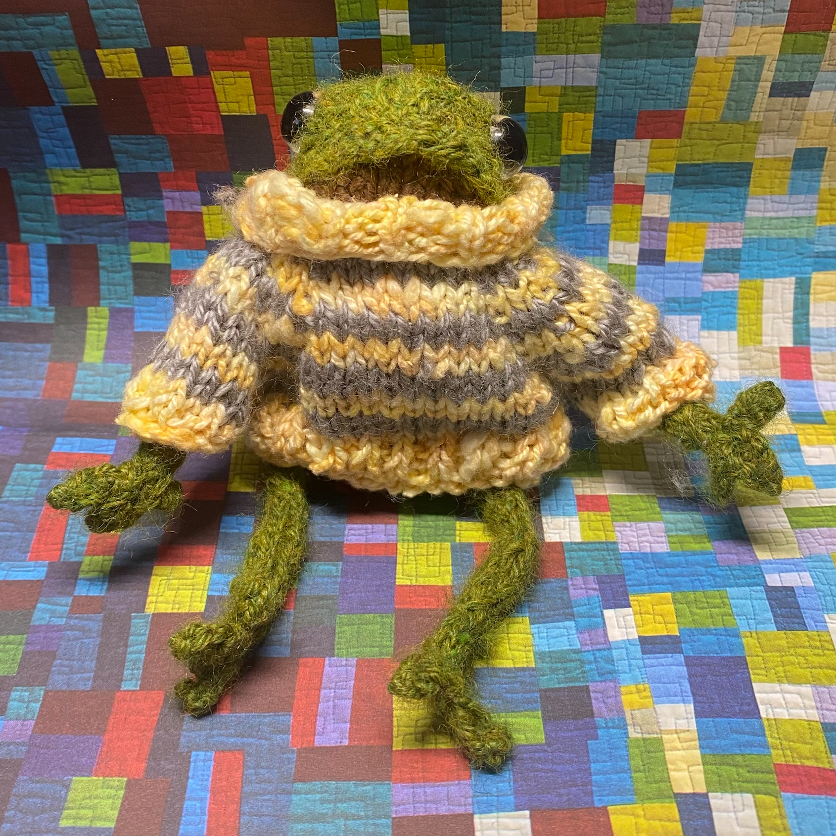 Trevor the toy frog is green with a brown belly and sits regally on a paper multicoloured quilted background. Trevor is wearing a tiny yet fashionable yellow and grey striped sweater and looks extra cozy.