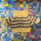 A yellow and grey striped knit sweater on a multicoloured paper quilt background. This sweater is for a toy frog with slight imperfections on the seam and left sleeve cuff. Despite its minor flaws, the sweater still features a fold-down collar and provides warmth and comfort to your toy frog.