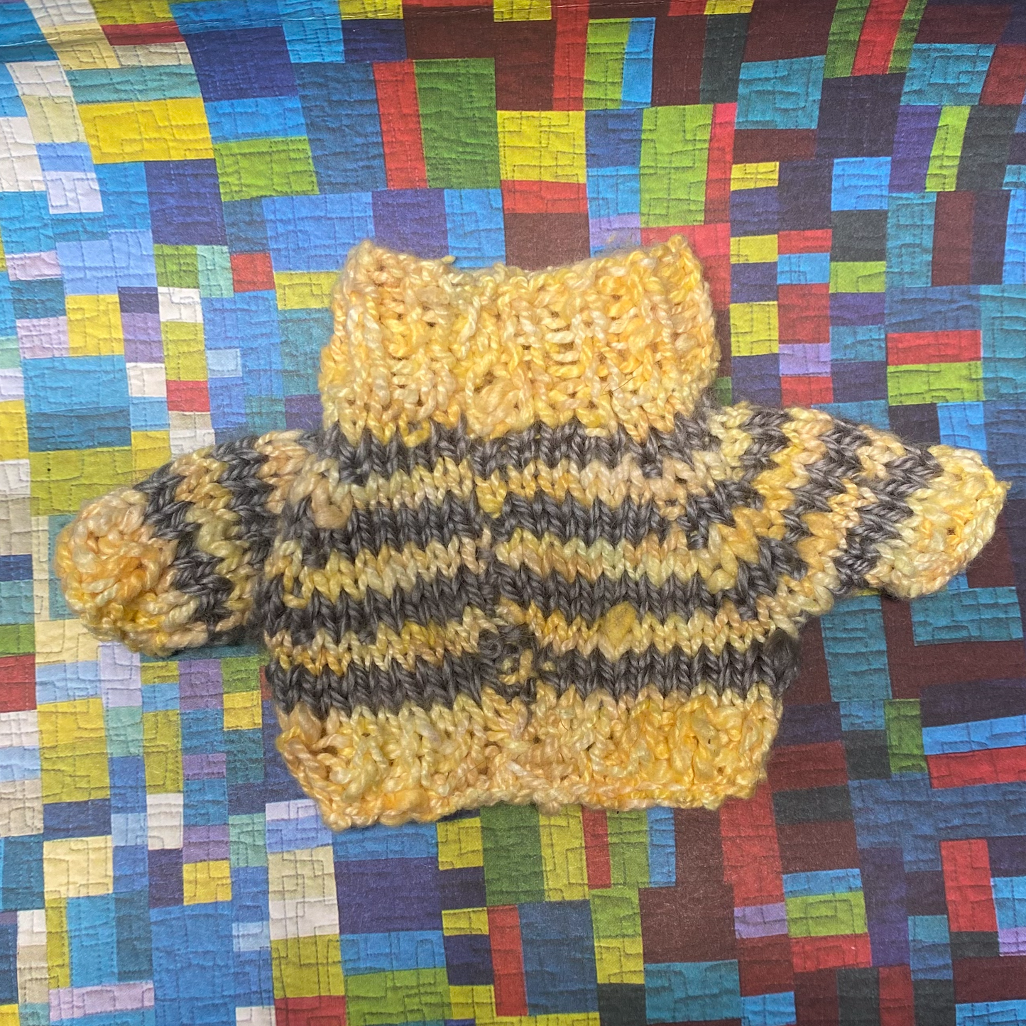 A yellow and grey striped knit sweater on a multicoloured paper quilt background. This sweater is for a toy frog with slight imperfections on the seam and left sleeve cuff. Despite its minor flaws, the sweater still features a fold-down collar and provides warmth and comfort to your toy frog.