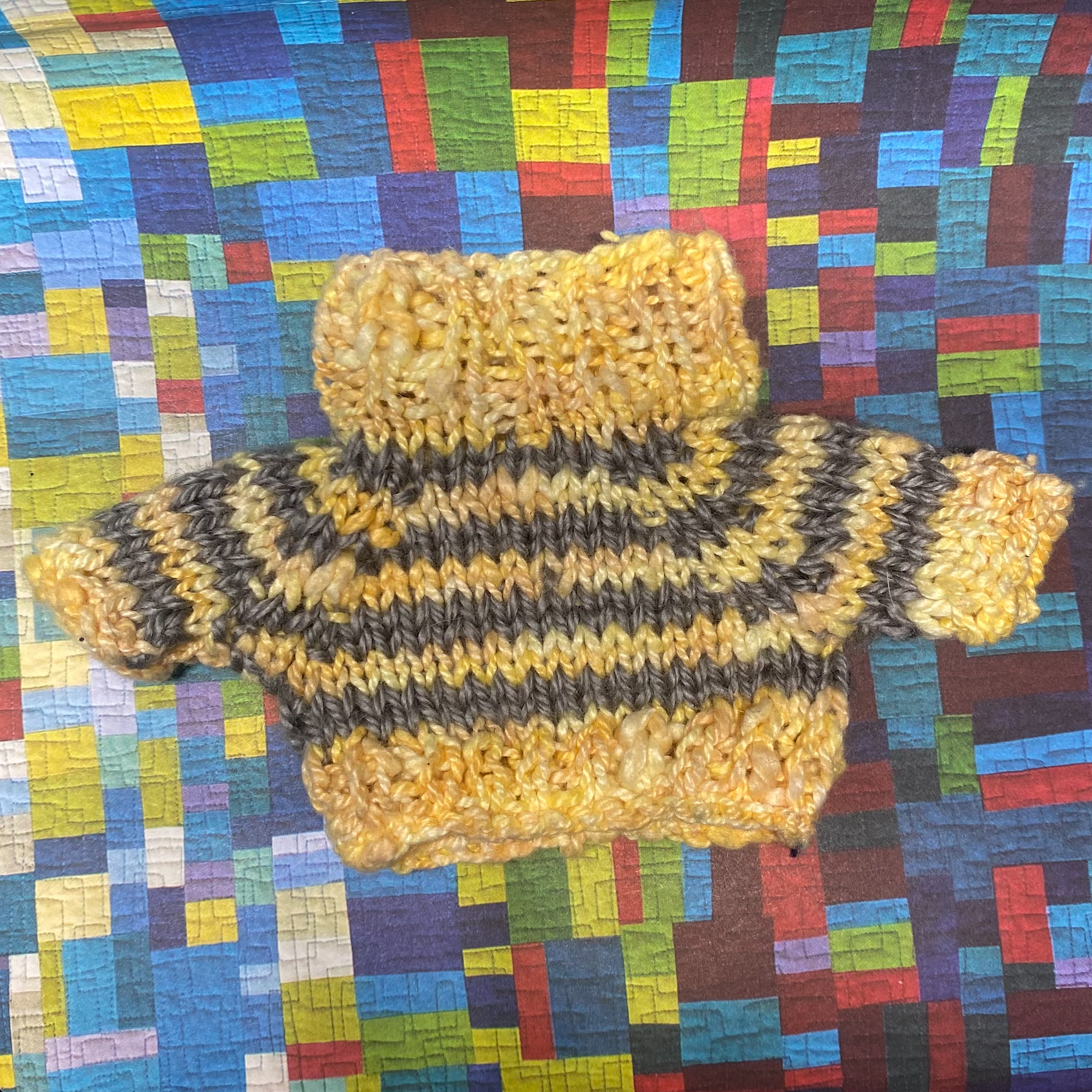 A yellow and grey striped knit sweater for a toy frog, positioned on a multicolored rectangular paper quilt background. The sweater features a fold-down collar and is designed to keep your toy frog cozy and stylish during the colder months.