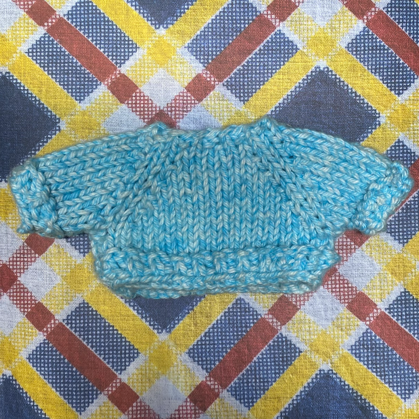 A cute knit frog sits on a plaid paper background, wearing a delightful blue sweater. The frog has large, round eyes and a friendly smile, and its arms hang down, emphasizing the coziness of the sweater. The sweater is light blue and features a detailed knit pattern.